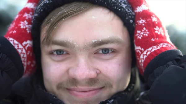 Portret van knappe, vrolijke man met snor gekleed in winterkleren met sneeuw op zijn gezicht. Close-up voor blonde man met blauwe ogen glimlachen en ontroerend zijn hoed in een zonnige winterdag. — Stockfoto