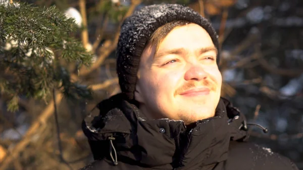 Portré, fiatal, mosolygós ember bajusz élvezi havazás téli erdőben, hunyorogva a szemét a fényes nap. Szép, boldog ember szereti hóesésben, a feje fölött a téli napsütésben. — Stock Fotó