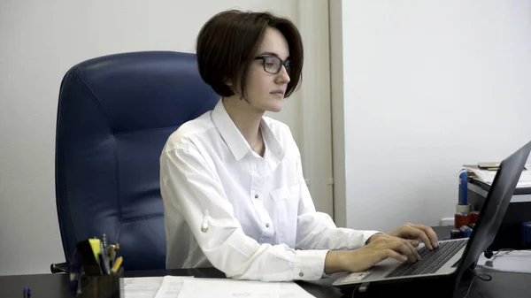 Молодая и красивая деловая женщина с короткими темными волосами в белой рубашке устала от работы в офисе. Молодая офисная леди печатает на компьютере, снимает очки и наклоняется назад в кресле . — стоковое фото
