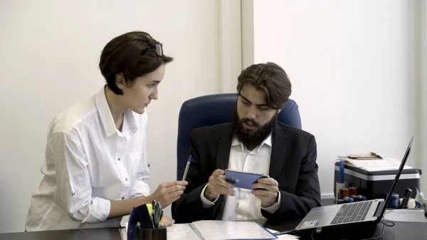 Kvinna sekreterare visar viktiga dokument till sin skäggiga chef som är upptagen med att spela smart telefon spel på kontoret, beroende av moderna enheter koncept. Affärsman tecknar papper ilsket. — Stockfoto