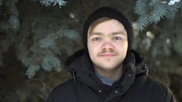 Porträt eines blonden Mannes mit Schnurrbart in warmer Kleidung vor grünem Fichtenhintergrund. Nahaufnahme für jungen Mann, der im Winterwald gegen Christbaum steht. — Stockfoto