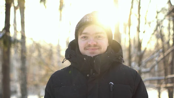 Nuori, hymyilevä mies auringon paisteessa talvimetsässä. Onnellinen mies talvivaatteissa hymyillen, katsellen suoraan kultaiseen auringonlaskuun lumisten puiden keskellä. . — kuvapankkivalokuva