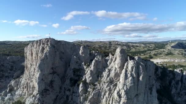 Cerca de los picos de montaña con una bandera y un campo verde detrás de ellos en el fondo azul, cielo nublado, la naturaleza por dron. Le dispararon. Aérea para acantilados impresionantes y prado verde en un día soleado de verano . — Vídeos de Stock