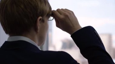 Pencereden dışarı bakan gözlüklü işadamının yakın çekim. Takım elbiseli genç işadamı gözlüklerini düzeltir, ofisten pencereden dışarı bakar