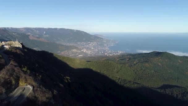 Vista aérea desde la alta montaña a una ciudad costera y empinada pendiente cubierta de árboles verdes. Le dispararon. Vista del paisaje marino de la ciudad costera en un valle verde sobre fondo azul, cielo claro . — Vídeo de stock