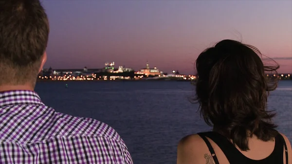 站在码头上的男人和女人。现场。从站在码头上的一男一女的背后特写镜头。从码头到另一边的闪亮城市的夜景与日落色的天空 — 图库照片