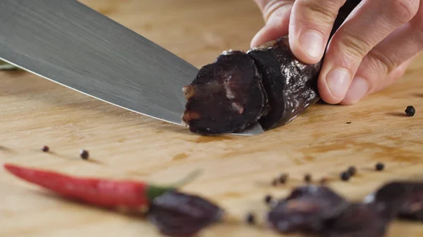 Primo piano di coltello taglia salsiccia. Scena. Primo piano del taglio di salame scuro sul tagliere di legno. Sale su sfondo di belle spezie — Foto Stock