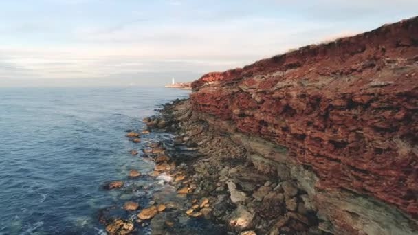 Rocky reef of shore. Shot. Top view of coastline of rocky seashore cliffs of red with holes and sticking stones. Beautiful blue sea comes and goes in waves — Stock Video