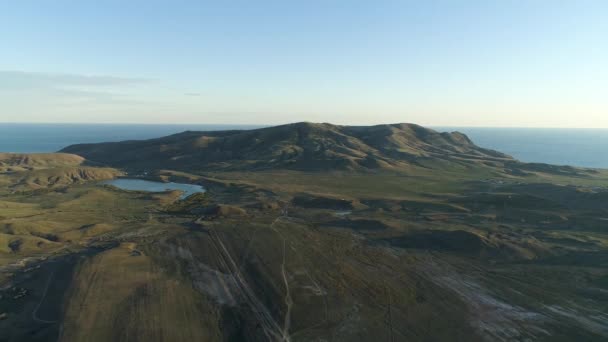 Vue panoramique aérienne du champ vert, de hautes montagnes près d'un petit lac sur fond de mer bleue et de ciel nuageux. Fusillade. Beau paysage avec de hautes collines verdoyantes et vallée près de la mer . — Video