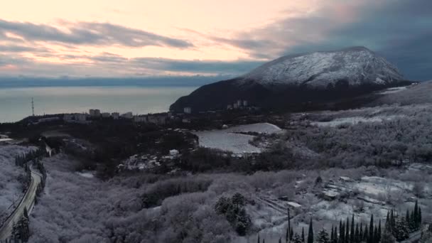 Vista panorâmica do aterro da cidade e montanha alta coberta de neve ao pôr-do-sol. Atingido. Aéreo para uma cidade costeira, árvores de inverno e campos, alta montanha coberta de neve contra o mar azul . — Vídeo de Stock