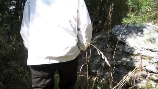 Vue arrière d'un homme en veste blanche marchant dans l'herbe haute sur le fond de la forêt, au ralenti. Des images. Forêt adulte dans le sentier forestier étroit en face des rochers . — Video