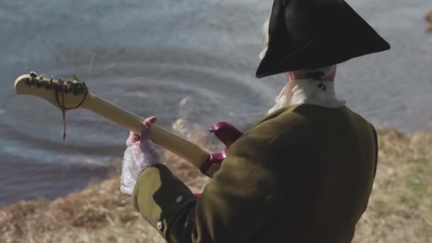 Achteraanzicht van een man in de oude ouderwetse kostuum en witte pruik met cocked hat gitaarspelen op de oever van een rivier. Voorraad. Man in de 17 eeuw kleding speelt gitaar met hangende algen op de rivieroever van een. — Stockvideo