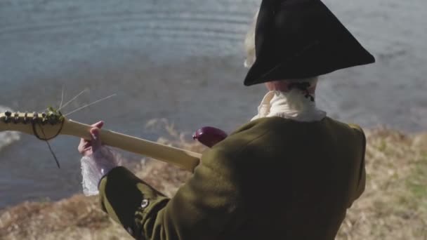 Rear view of a man in old fashioned costume and white wig with cocked hat playing guitar on a river shore. Stock. Man in 17 century clothes plays electric guitar with hanging algae on a river bank. — Stock Video