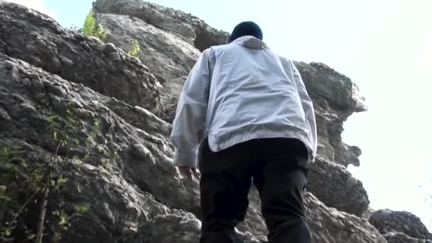 Close up of male climbing high mountain on the background of trees and blue sky, view from below. Footage. Rear view of a man standing on rocks, travel, sporty lifestyle concept. — Stock Video