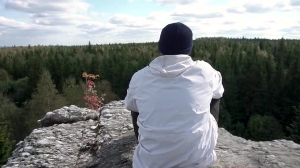 Zadní pohled na horský turista při pohledu na podzimní krajinu lesní a zamračená obloha. Muž sedí na vrcholu vysoké skály při pohledu na vrcholky stromů. — Stock video