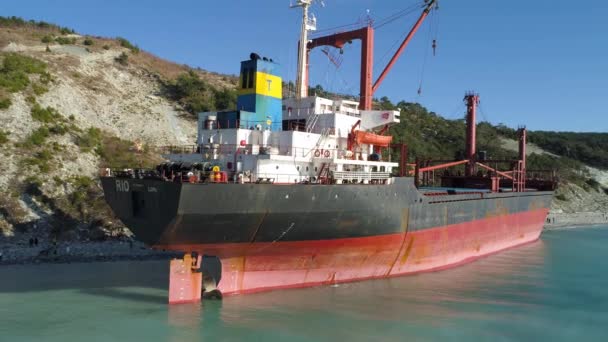 Aerial for an empty industrial ship moored near sea shore with many people walking on a beach. Maritime cargo vessel standing near green trees slope in a summer sunny day. — Stock Video