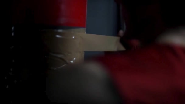 Rear view of a man preparing his boxing pear by wrapping it with sellotape in the dark room, sport concept. Fighter wrapping punching bag with brown sticky tape. — Stock Video