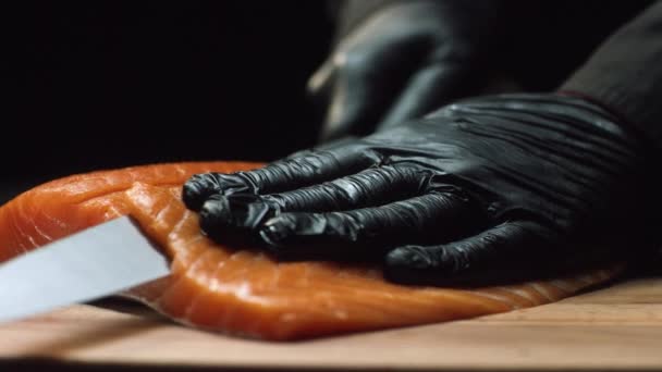 Chef de cerca con trozo de salmón rojo. El chef profesional en guantes negros corta trozo de salmón fresco en rodajas finas con un cuchillo afilado — Vídeos de Stock