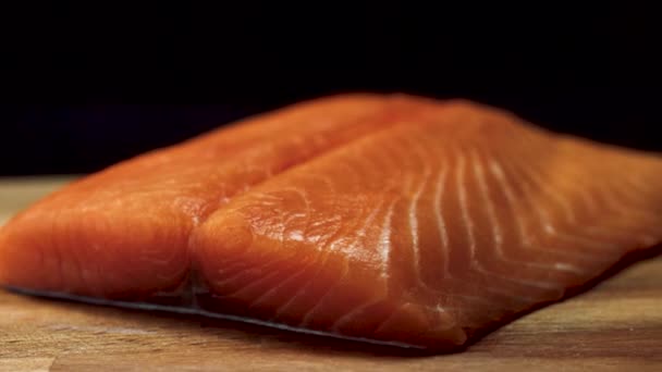 Primer plano del jugoso trozo de salmón. Jugosa rebanada fresca y roja de carne de salmón acostada sobre tabla de madera sobre fondo negro — Vídeos de Stock