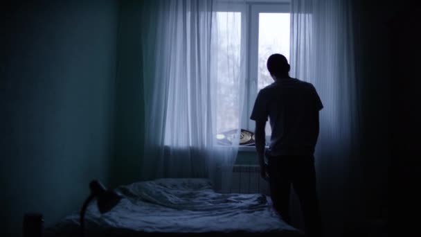 Un hombre mirando por la ventana de su habitación. Vista desde atrás de un joven cansado parado en la ventana por la noche — Vídeos de Stock