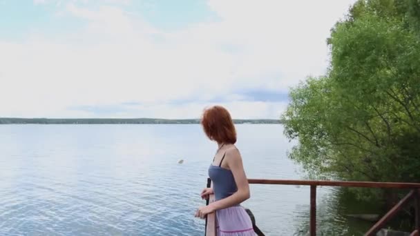 Attractive young red-haired woman on pier in summer. Beautiful young woman in long skirt and t-shirt on village pier enjoys summer beautiful weather — Stock Video