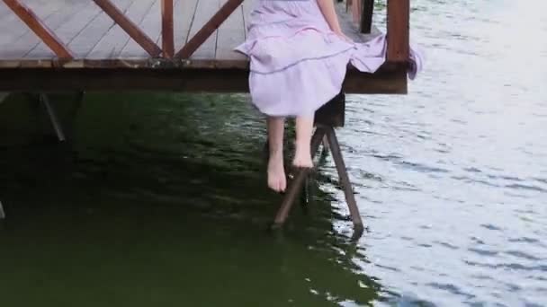 Aantrekkelijke jonge vrouw zittend op de pier in de zomer. Roodharige heldere mooie jonge vrouw in lange rok zittend op de pier op achtergrond van groene vegetatie — Stockvideo