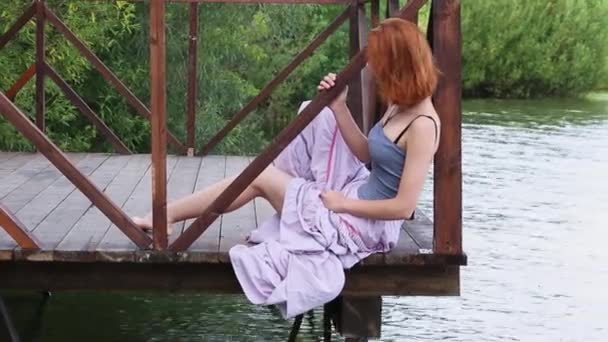 Aantrekkelijke jonge vrouw zittend op de pier in de zomer. Roodharige heldere mooie jonge vrouw in lange rok zittend op de pier op achtergrond van groene vegetatie — Stockvideo