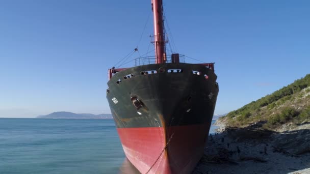 Aerial for tug boat moored on the ocean shore near the hill with green trees on blue sky background. Shot. Red industrial ship near forested slope and people walking around. — Stock Video