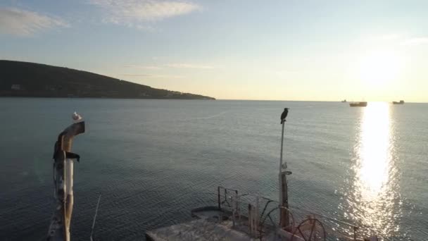 Aérea para parte de un barco con un pájaro sentado en un pequeño poste de madera con cielo azul, nublado y fondo verde de montaña. Le dispararon. El borde del barco y el reflejo de la luz solar en la superficie del agua . — Vídeo de stock