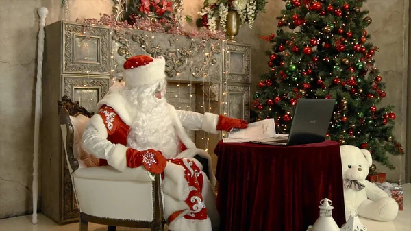 Le Père Noël moderne lit des lettres avec un ordinateur portable. Clip. Le Père Noël lit une lettre dans une pièce avec un sapin de Noël — Photo