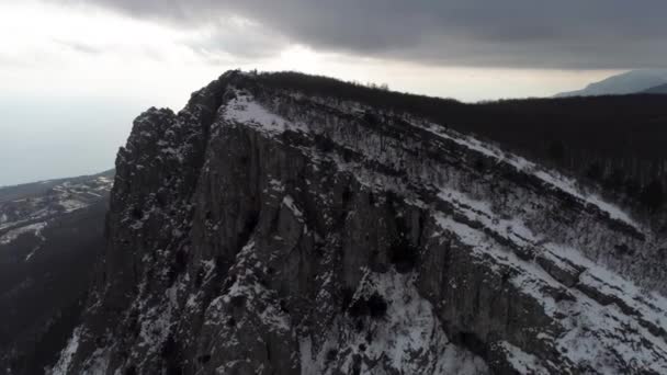 La pente du haut massif rocheux recouvert de pins dans des nuages brumeux. Fusillade. Aérien pour le versant boisé de la montagne dans les nuages lourds et gris et le littoral avec la petite ville . — Video