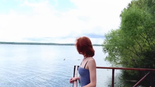Mujer joven, delgada, pelirroja, de pie en una cubierta junto al agua, mirando a la distancia. Chica reflexiva brillante de pie en el muelle cerca del lago . — Vídeos de Stock