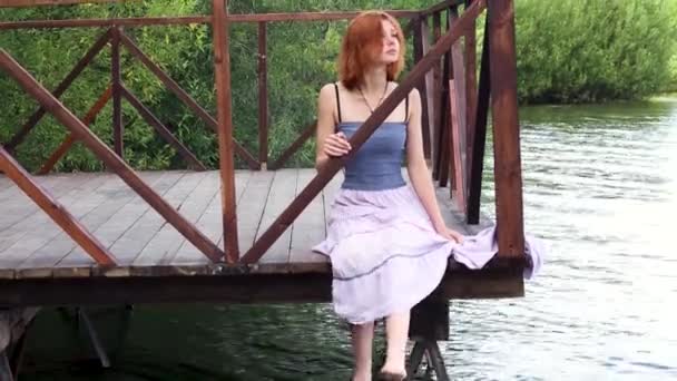 Молода, струнка, руда волохата жінка сидить на палубі біля води, дивлячись на відстань. Яскрава вдумлива дівчина сидить на пірсі в довгій спідниці біля озера . — стокове відео