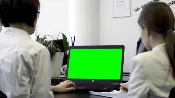 Young women working in office, sitting in chairs in front of computer with green screen, rear view. Two girls co workers looking at computer monitor with chroma key over white wall background.