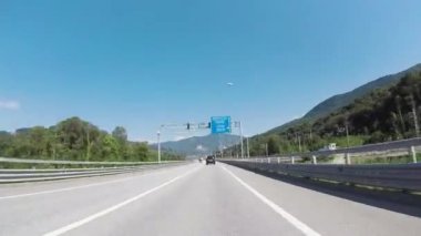 Yolun bir güneşli, ağaçların arasında karayolu üzerinde yaz günü hareketli araba penceresinden görünümü. Sahne. Bulutlu, mavi gökyüzü ve ormanlık dağlar karşı yolda sürüş araç.