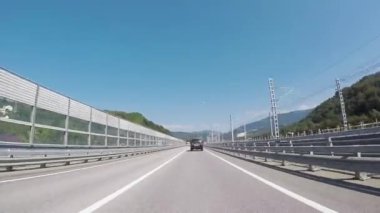 Yolun bir güneşli, ağaçların arasında karayolu üzerinde yaz günü hareketli araba penceresinden görünümü. Sahne. Bulutlu, mavi gökyüzü ve ormanlık dağlar karşı yolda sürüş araç.