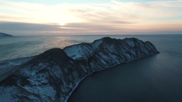 Letecký pohled na skalnatém mysu pokryty sněhem v tmavé mořské vodě proti zamračená obloha při východu slunce. Střela. Krásné mořské krajiny. — Stock video