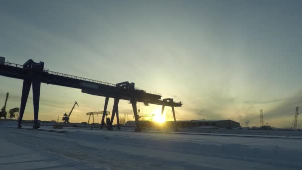 A large gantry crane standing still on the rail while automobile cranes worki behind it, time lapse. Gantry crane in the industrial zone in winter morning on rising sun background. — Stock Video