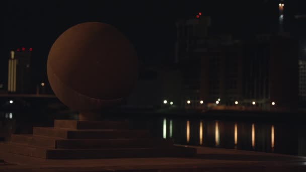 Escultura de pelota en la ciudad por la noche. Escultura esférica de piedra en muelle sobre fondo de luces de la ciudad reflejadas en el agua por la noche. Escultura geométrica simple en paisaje de ciudad — Vídeos de Stock