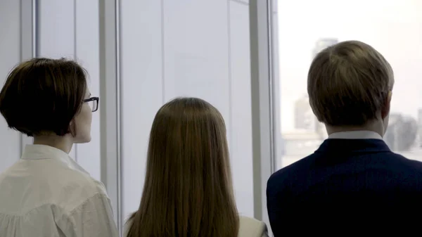 Rückansicht einer Unternehmensgruppe, die in einer Reihe steht und durch ein Fenster blickt. zwei weibliche und männliche Mitarbeiter stehen zusammen im Büro in der Nähe des großen Fensters und schauen auf die Stadt. — Stockfoto