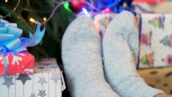 Feet in knitted socks near New Year presents on Christmas tree background, winter holidays and home comfort concept. Close up for cozy and warm white socks near wrapped gifts over decorated spruce. — Stock Photo, Image
