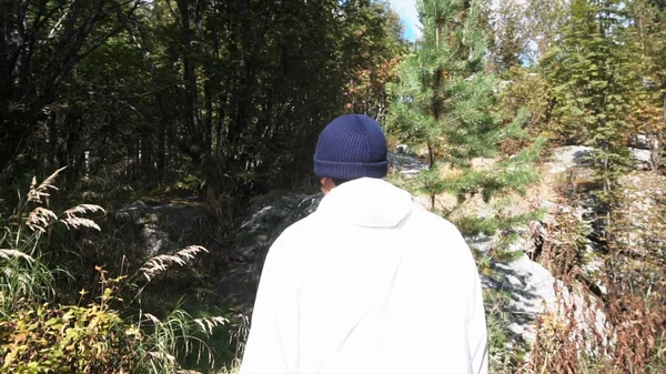 Οπίσθια όψη ενός άνδρα στο λευκό σακάκι με τα πόδια σε ψηλό χορτάρι στο δάσος φόντο, αργή κίνηση. Μήκος σε πόδηα. Ενηλίκων δασοφύλακας στο στενό δασικό δρόμο μπροστά από τα βράχια. — Φωτογραφία Αρχείου