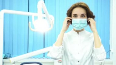 Bir diş hekimi diş Kliniği kameraya bakarak maskesi üzerine koyarak portresi. Kadın diş hekimi asistanı ayakta silah ile ameliyat maskesi yüzünde geçti..