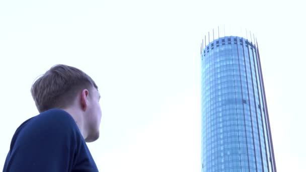 Вид сзади на человека, стоящего и смотрящего на высокую, современную башню со стеклянным фасадом и жеванием. Мужчина в черном кардигане смотрит на здание современного бизнеса на ярком фоне неба . — стоковое видео