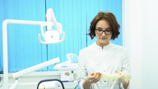 Mladá žena doktor nosí brýle a v bílém plášti, na bílých lékařských glovesin zubní klinika kabinetu. Přísná žena zubař klade na rukavice a hledí do kamery, příprava. — Stock video