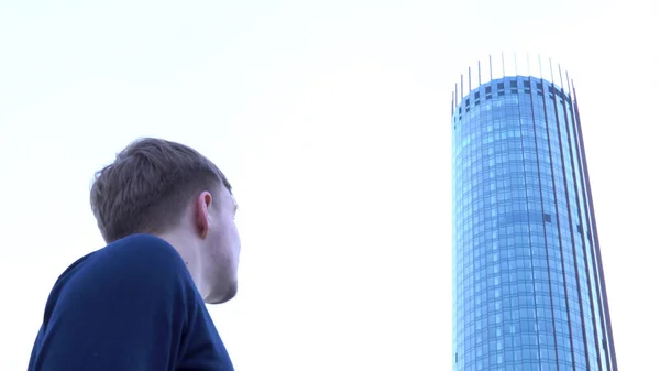 Vista traseira de um homem de pé e olhando para torre alta e moderna com fachada de vidro e mastigação. Masculino em cardigan preto olhando para o moderno edifício busines no fundo do céu brilhante . — Fotografia de Stock