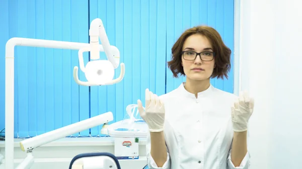 Νεαρή γυναίκα γιατρός φοράει γυαλιά και ένα άσπρο παλτό, βάζοντας στο λευκό ιατρικό glovesin οδοντιατρική κλινική Υπουργικού Συμβουλίου. Μια αυστηρή γυναικεία οδοντίατρος βάζει γάντια και φαίνεται στην κάμερα, ετοιμάζεται. — Φωτογραφία Αρχείου
