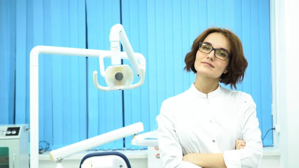 Portret van mooie jonge vrouw tandarts staande armen gekruist in de tandheelkundige kliniek kabinet. Zeker dokter in glazen op zoek direct naar de camera, geneeskunde concept. — Stockfoto