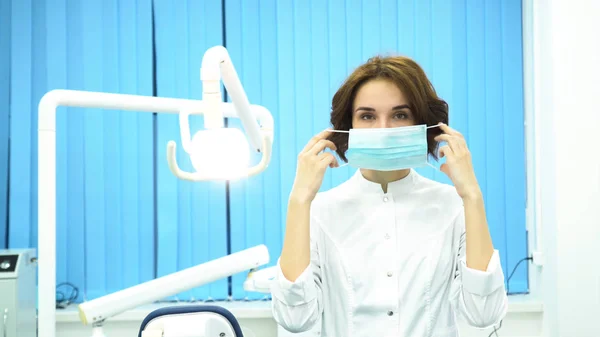 Portret van een tandarts het masker kijken camera bij de tandheelkundige kliniek te zetten. Vrouwelijk tandarts assistent staande armen gekruist met chirurgische masker op haar gezicht. — Stockfoto