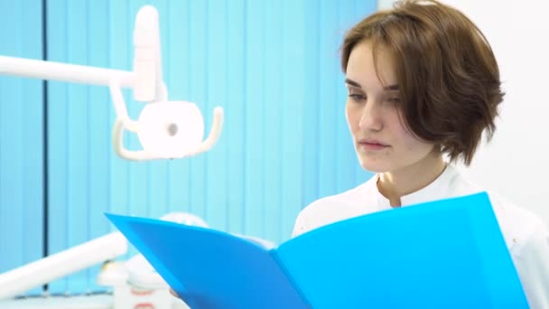 Doctora en gabinete de dentista leyendo notas de pacientes frente a equipos médicos, concepto de atención médica. Mujer dentista leyendo archivos en carpeta azul, pasando páginas . — Vídeo de stock